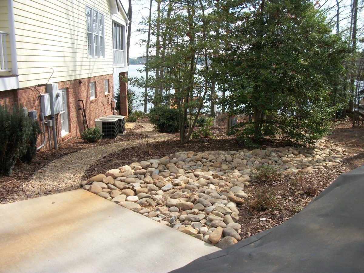 Front yard landscape with stone drainage bed, shrubs, and house siding beside a driveway