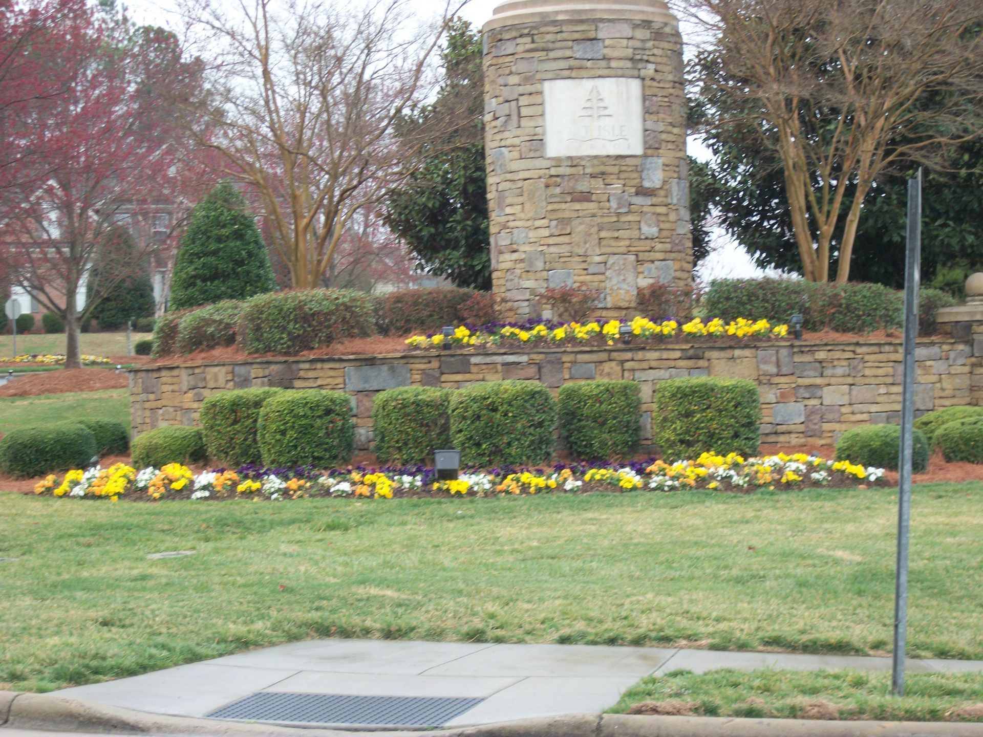 Landscaped memorial with stone monument, yellow flowers, trimmed hedges, and grass beside a sidewalk