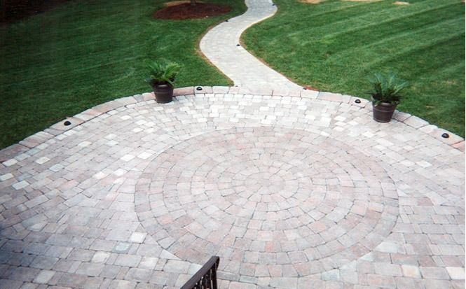 Curved stone patio bordered by green lawn with potted plants and a winding path beyond