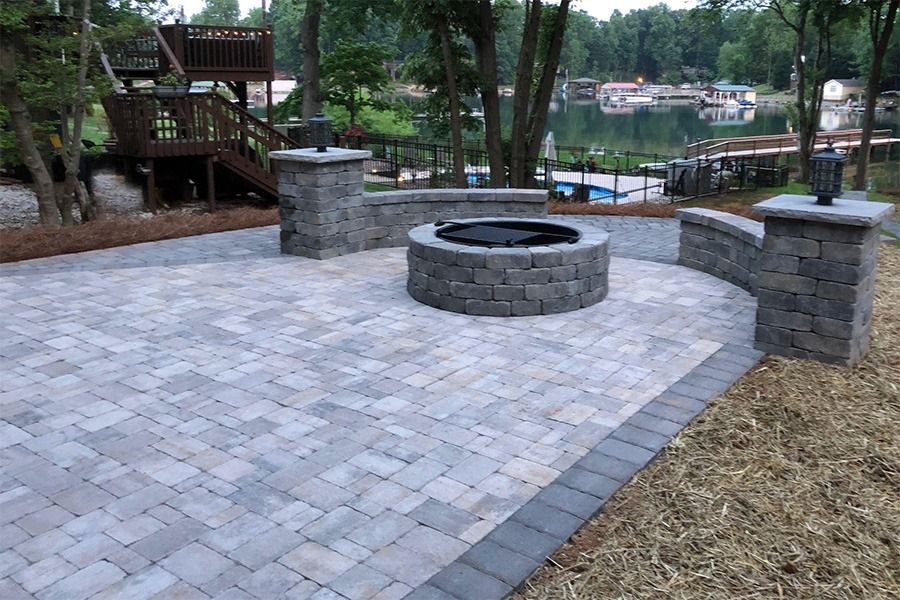 Stone fire pit on a paved patio with stone pillars, wooden deck, and wooded lakefront view