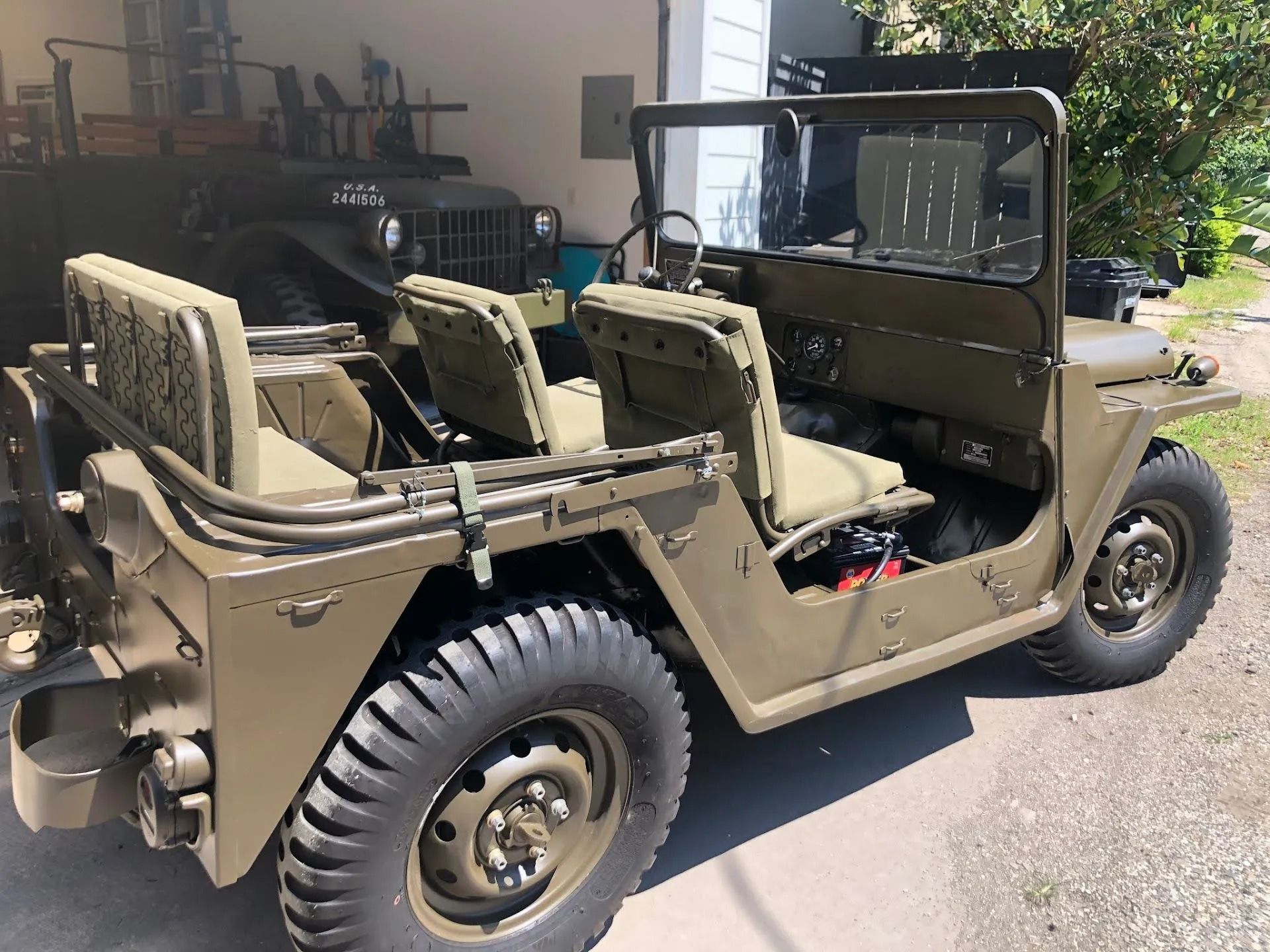 Olive drab military jeep parked outdoors beside a building, with tan seats and folded windshield.