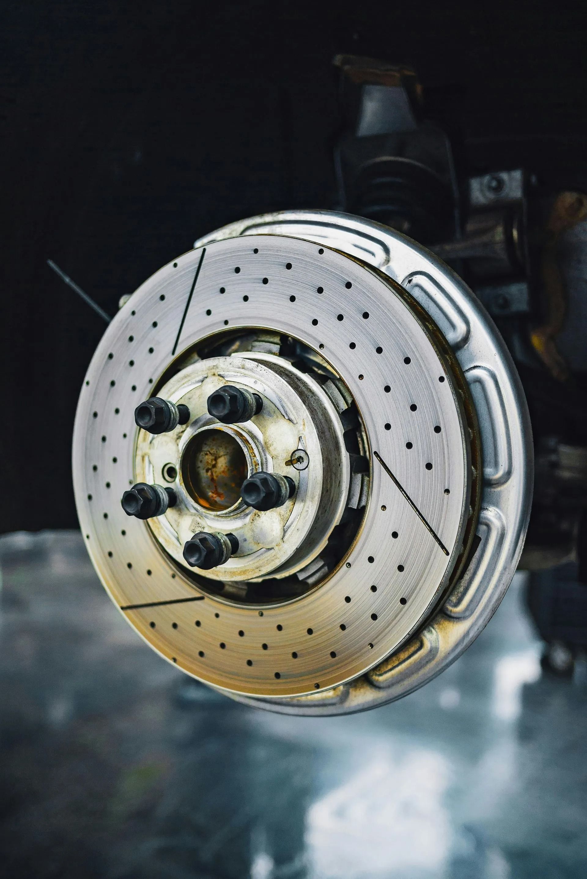 Close-up of a perforated car brake rotor and hub assembly in a dim garage, with smoke or mist nearby