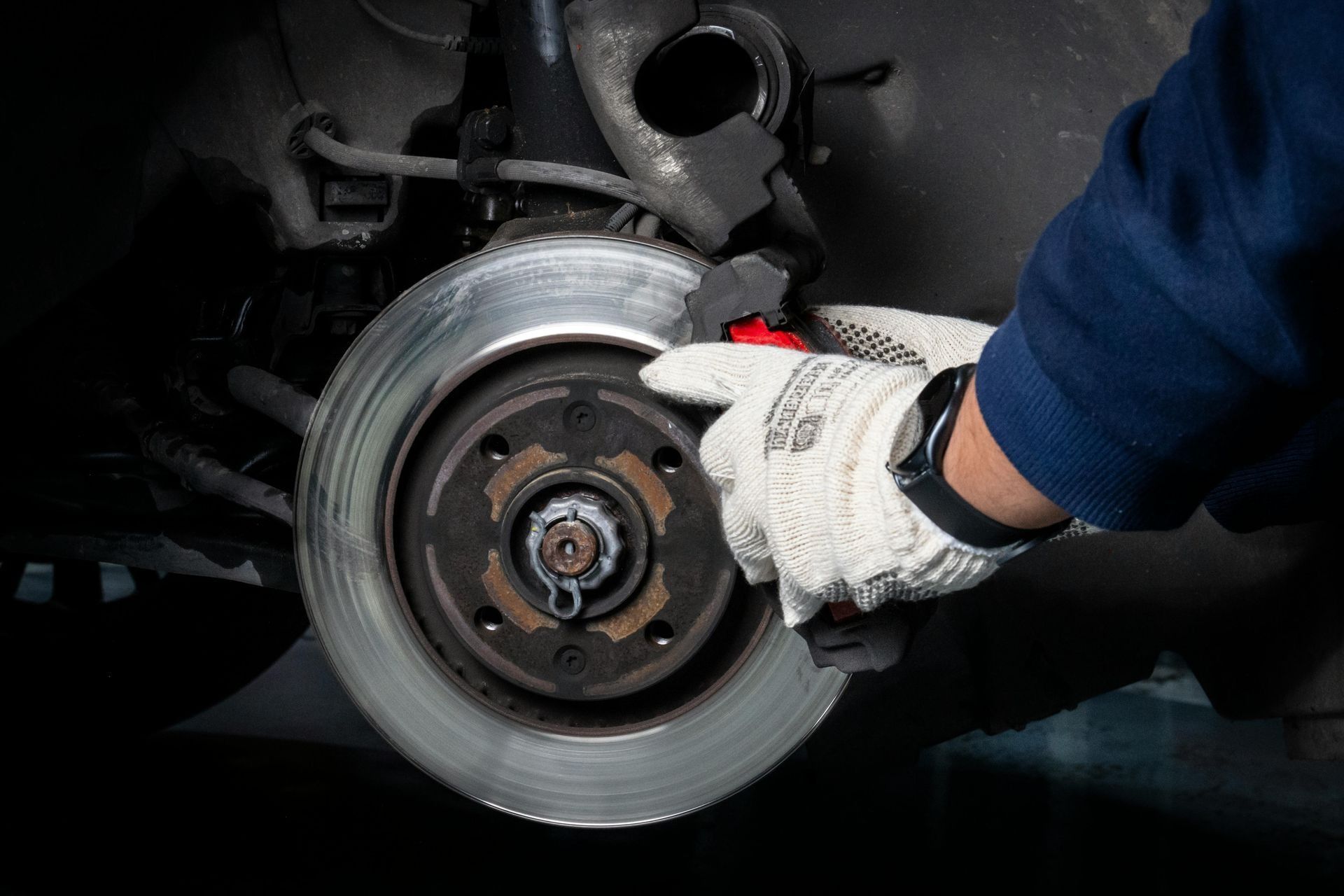 Gloved mechanic inspecting a car brake rotor with a hand tool in a dark garage