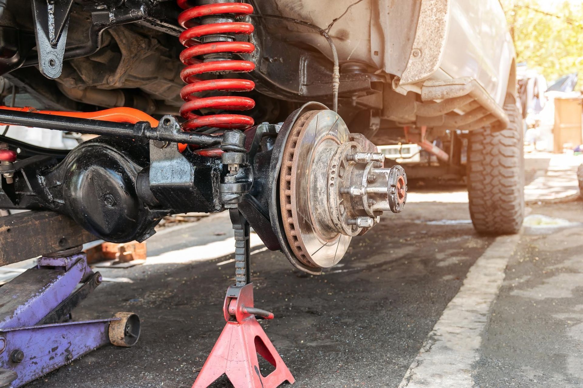 Car underside on jack with exposed brake rotor and red suspension spring during repair