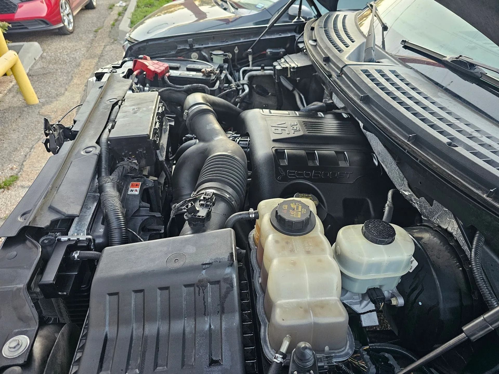 Car engine bay with hoses, fluid reservoirs, and black plastic covers, viewed from above