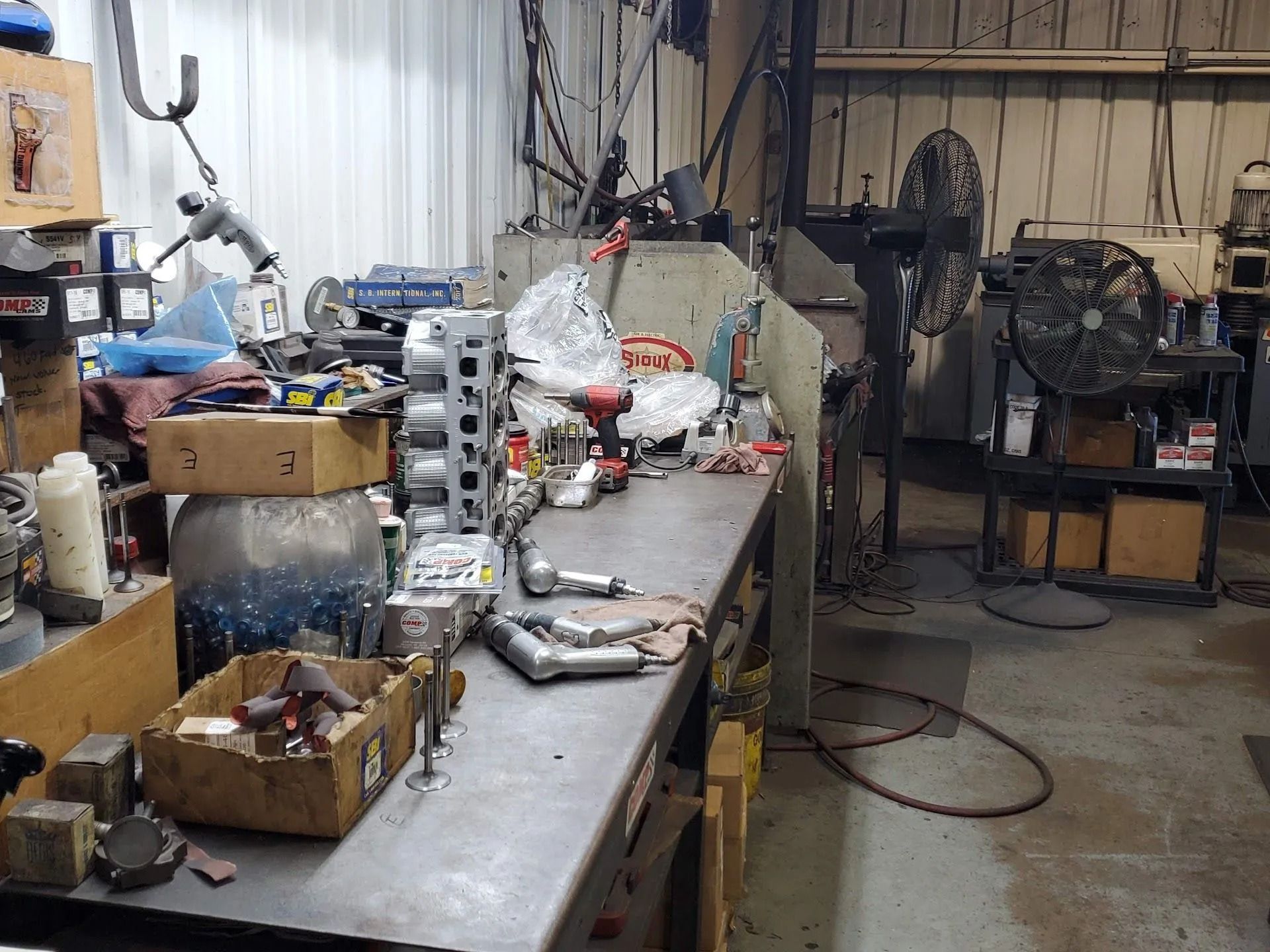 Cluttered workshop with workbench, tools, boxes, and machinery in a dim industrial space