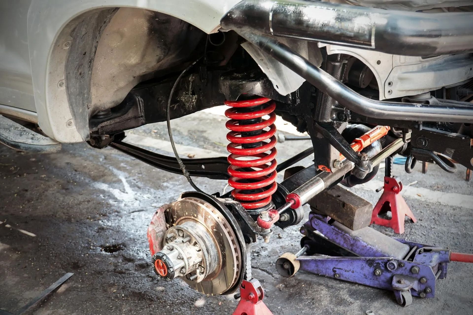 Rear car suspension and wheel hub under repair, with red coil spring and jack stands in a garage