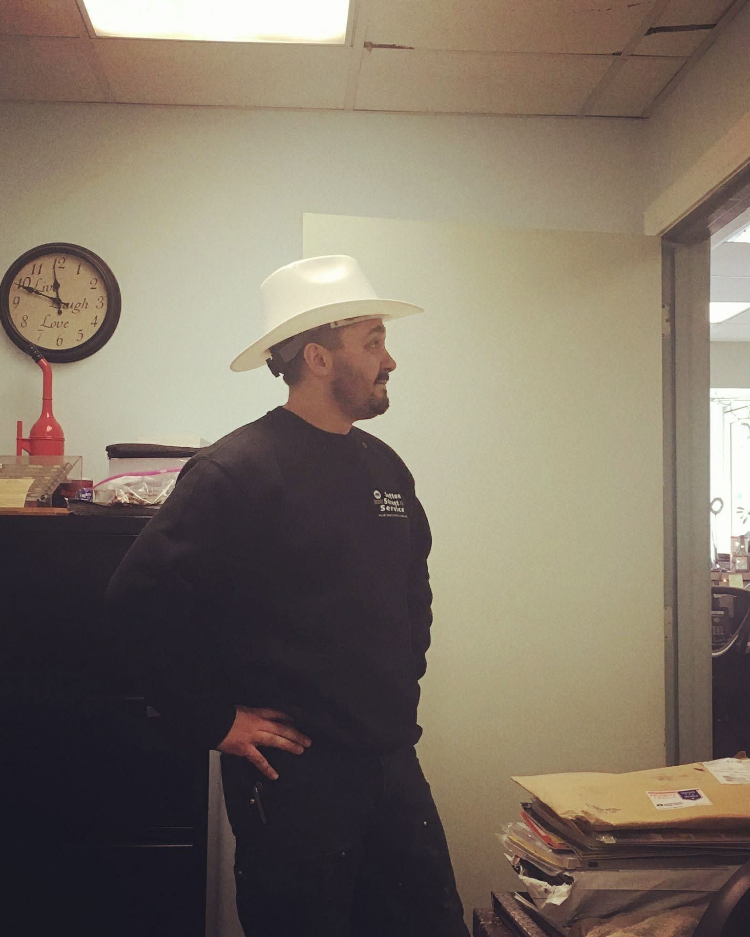 Man in a white hat stands in an office beside a desk with stacked papers.