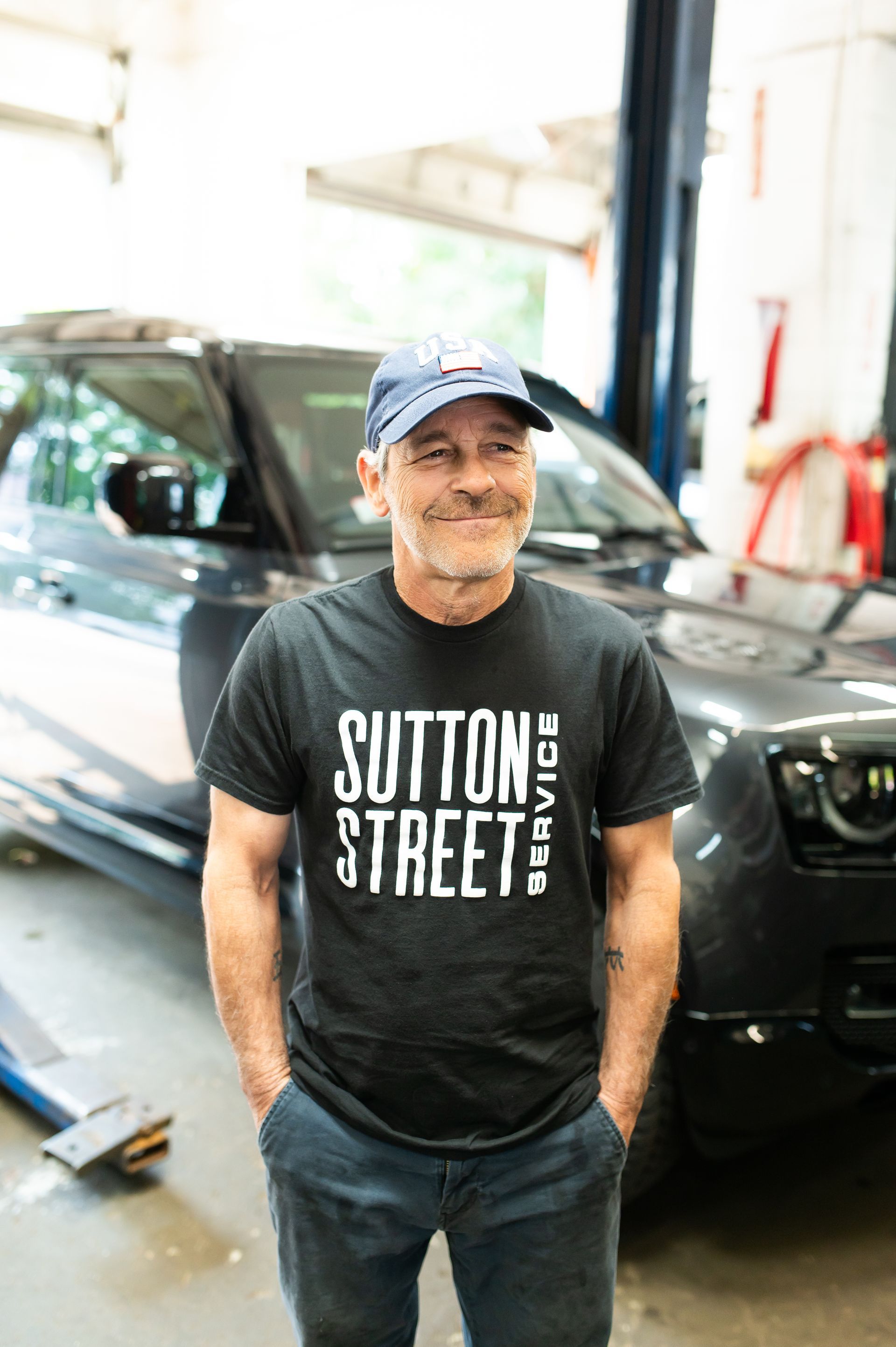 Man standing in a garage beside a black SUV, wearing a blue cap and a black Sutton Street T-shirt.