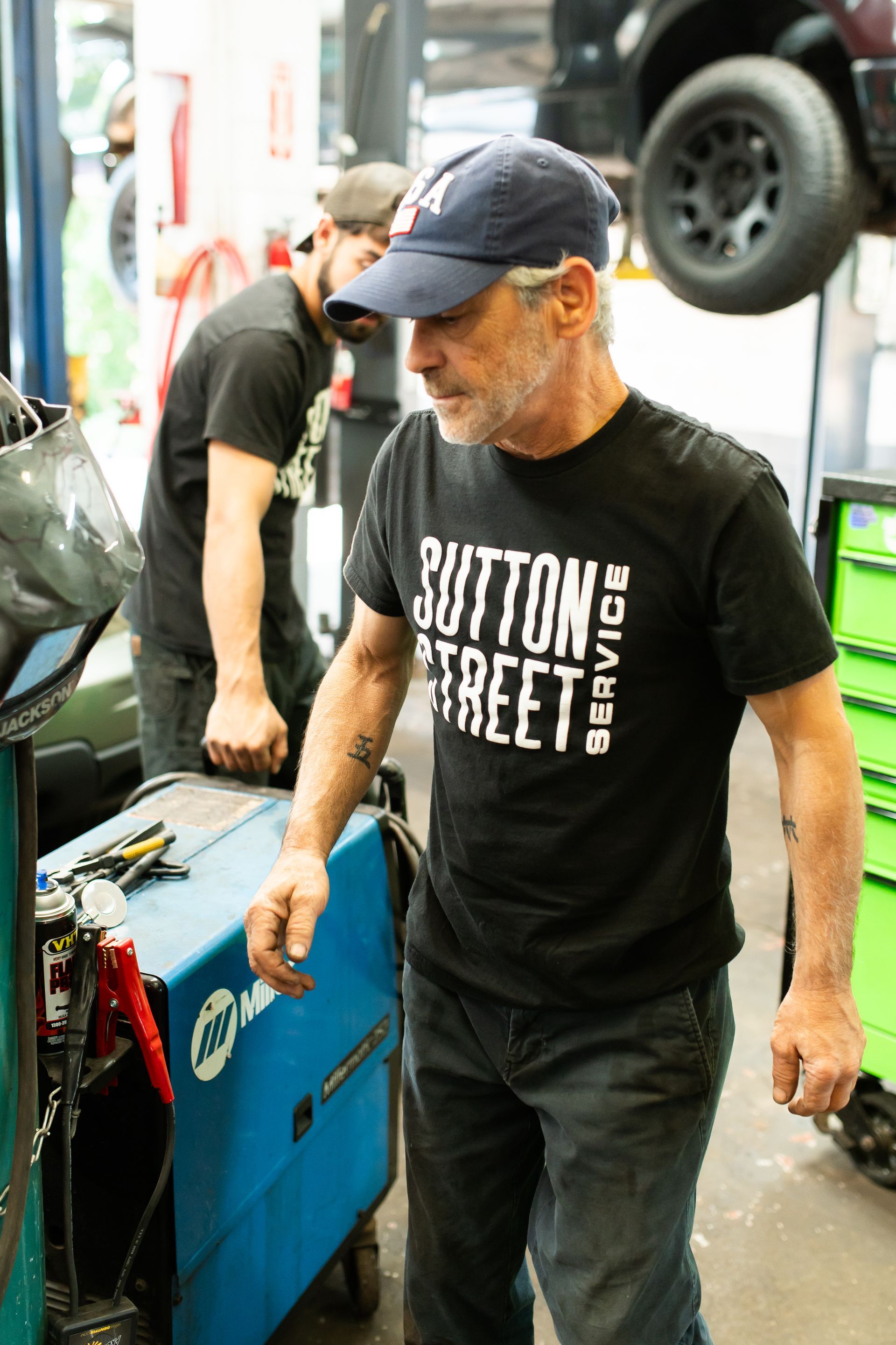 Man in black “Button Street” shirt and cap standing beside a blue cart in a workshop гараж-like setting