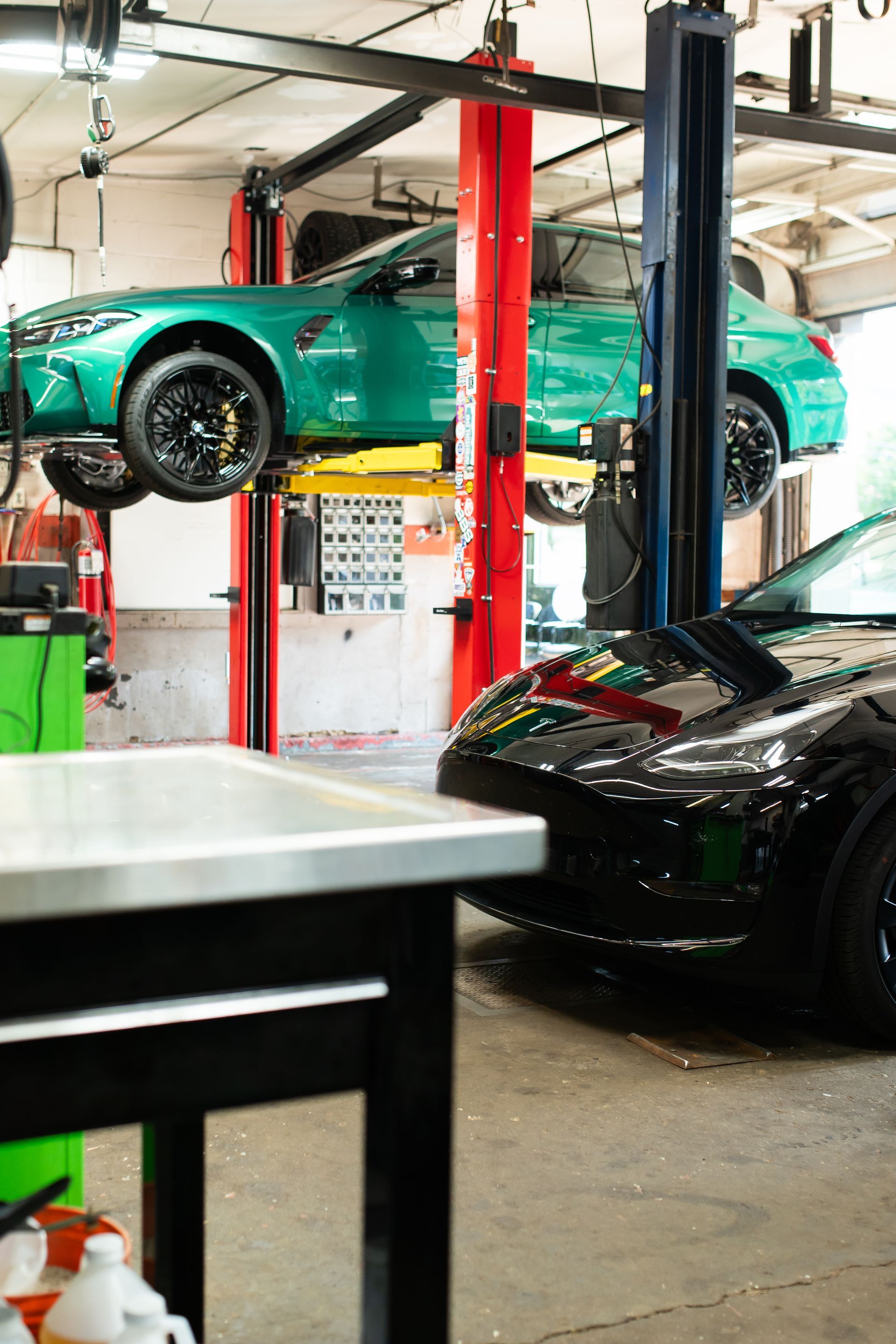Green sports car lifted on a hydraulic hoist in an auto repair shop, with a black car below.