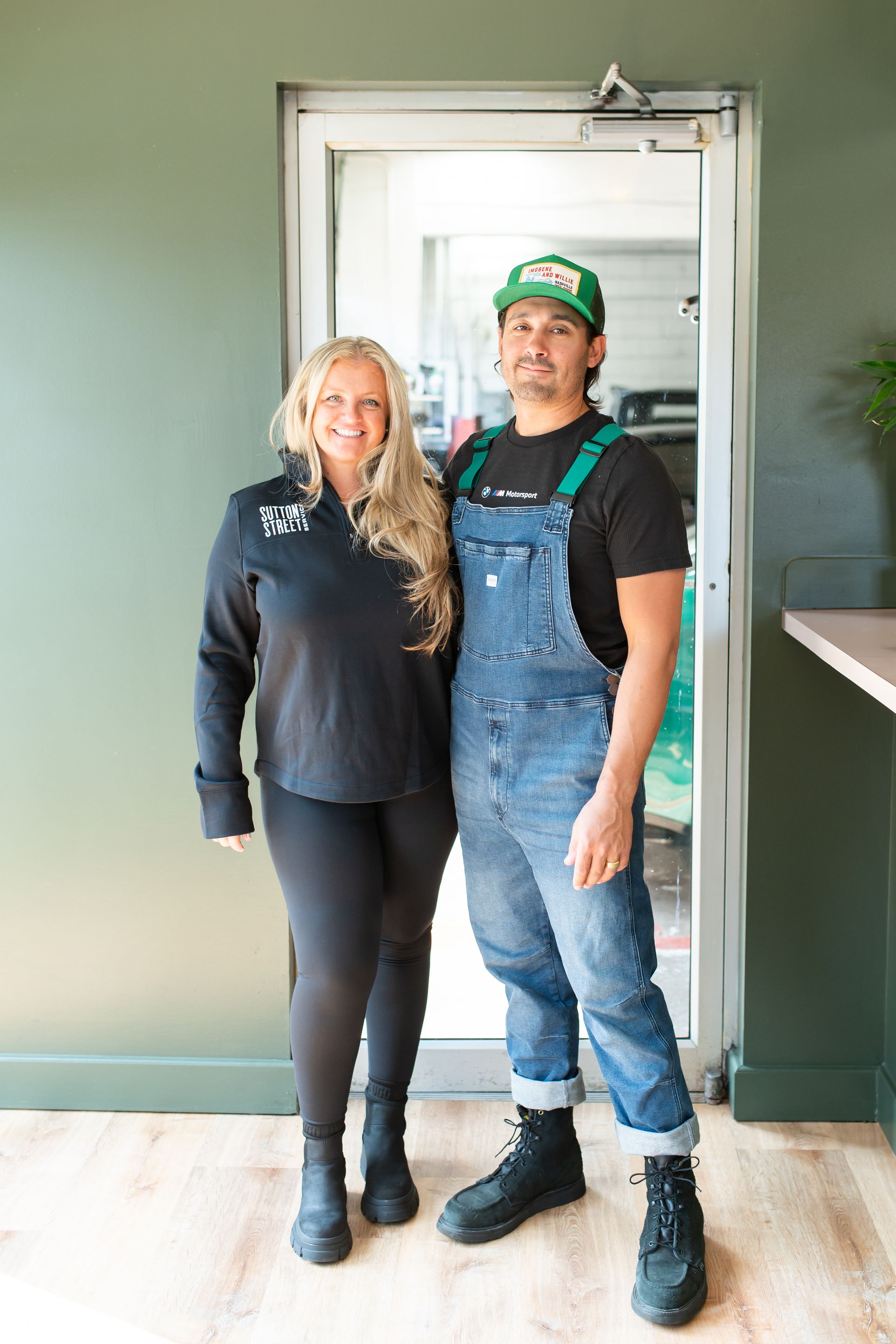 Two people standing in a doorway, one in black and one in denim overalls, against a green wall.