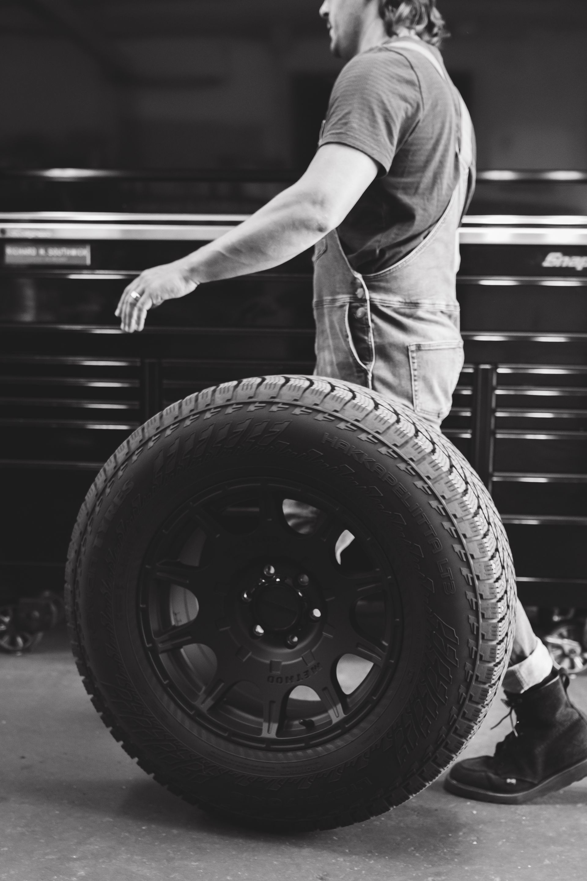 Person in a garage rolling a large off-road tire beside a workbench.