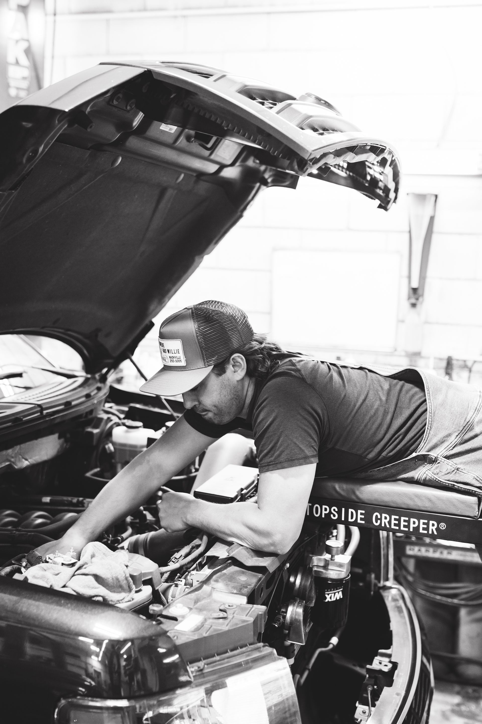 Mechanic working under a car hood in a garage, reaching into the engine bay.