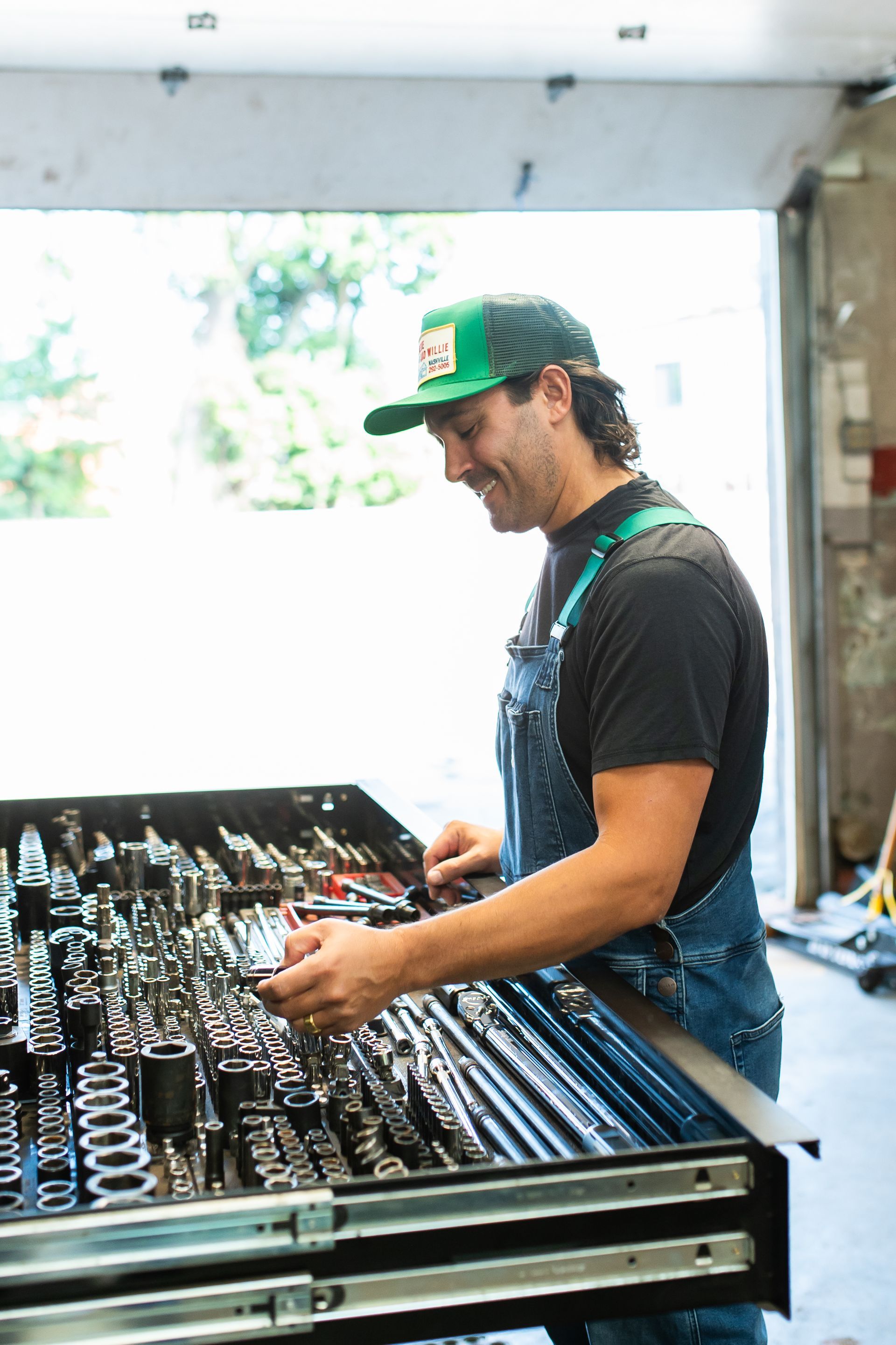 Worker in a green cap and black shirt adjusting tools at a workshop bench with metal parts.