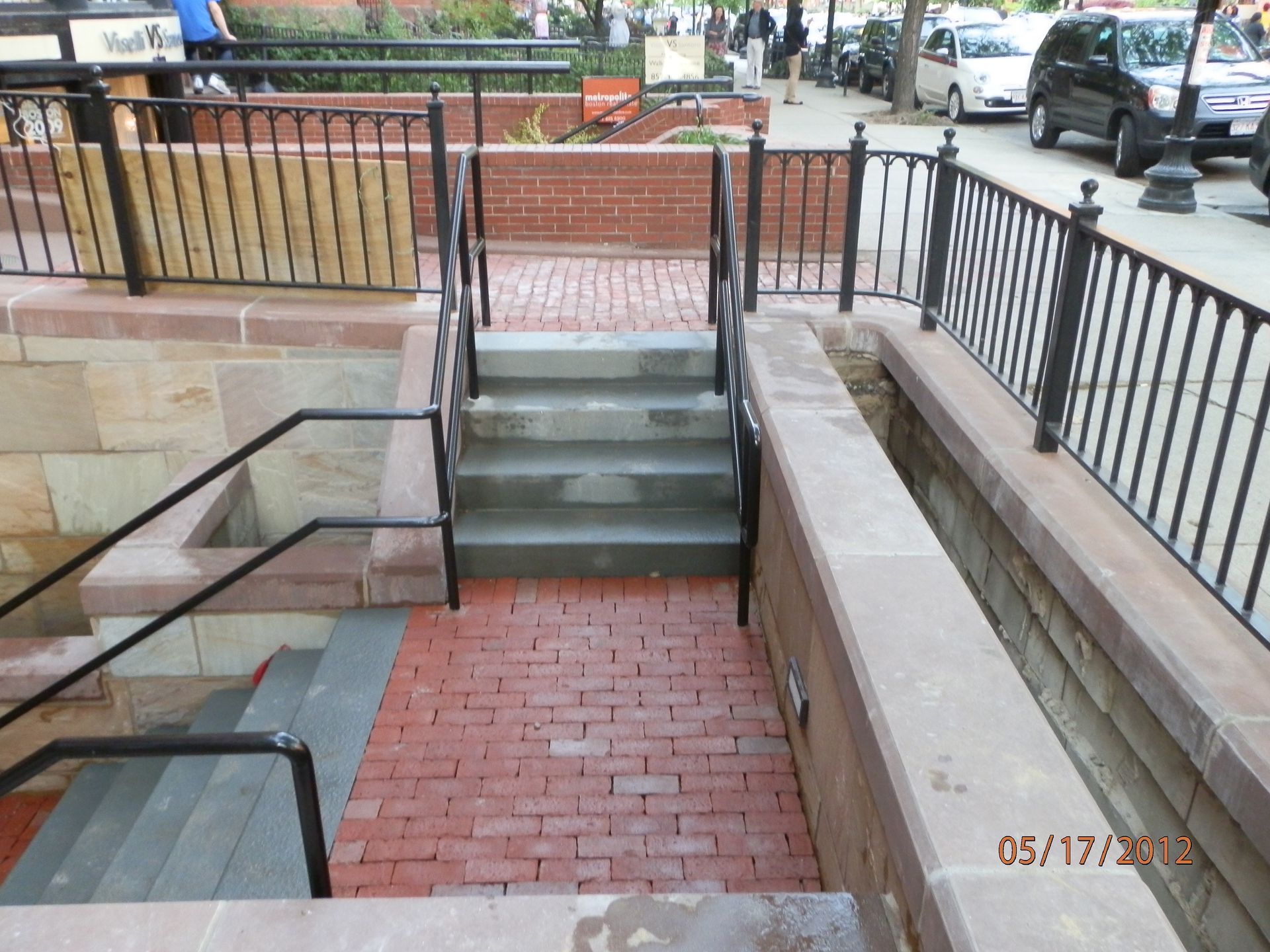 Brick stairwell and ramp entrance beside a sidewalk, with black railings and stone walls.
