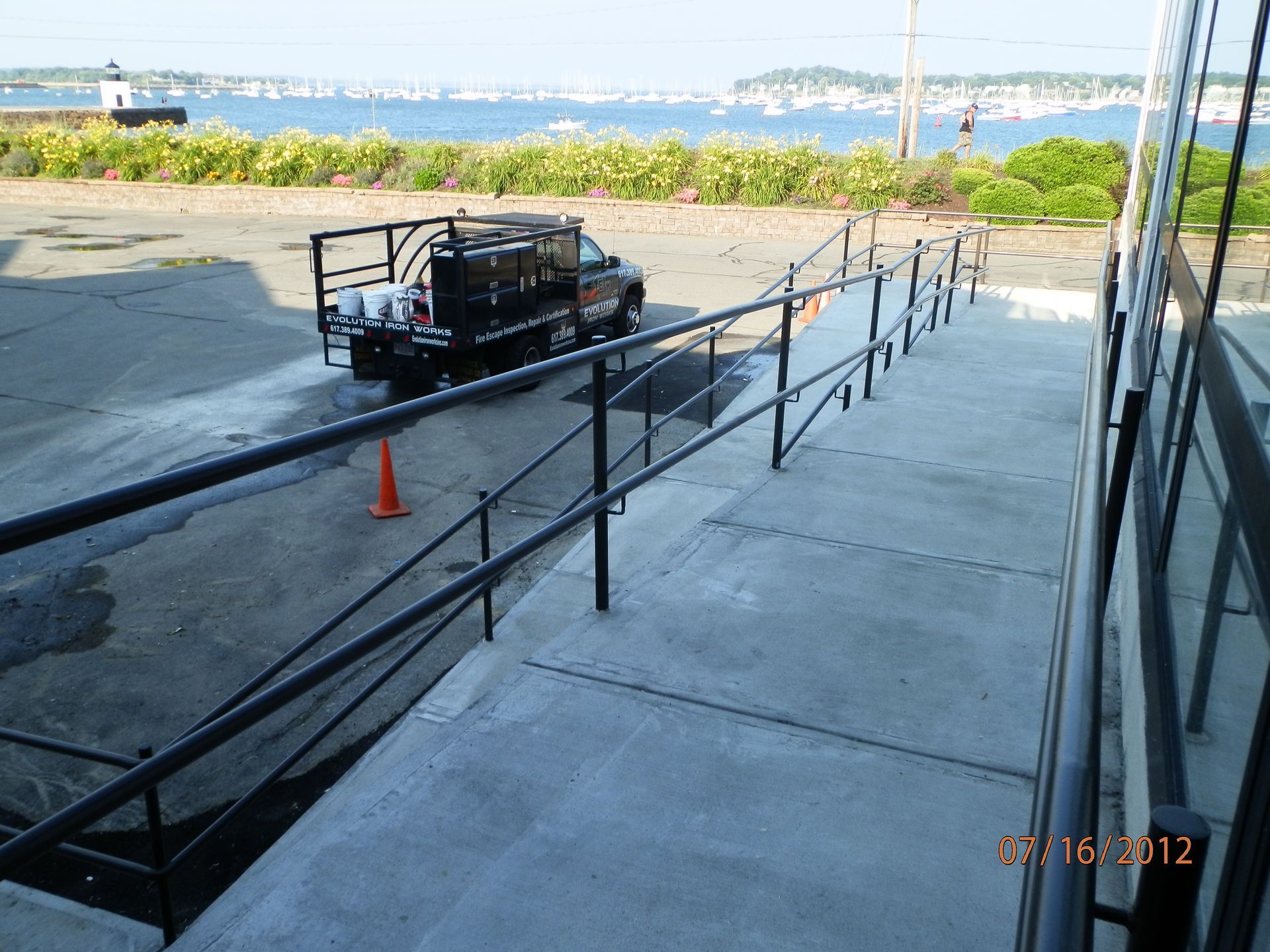 Outdoor ramp with black railings beside a waterfront, a utility cart and orange cone near the top.