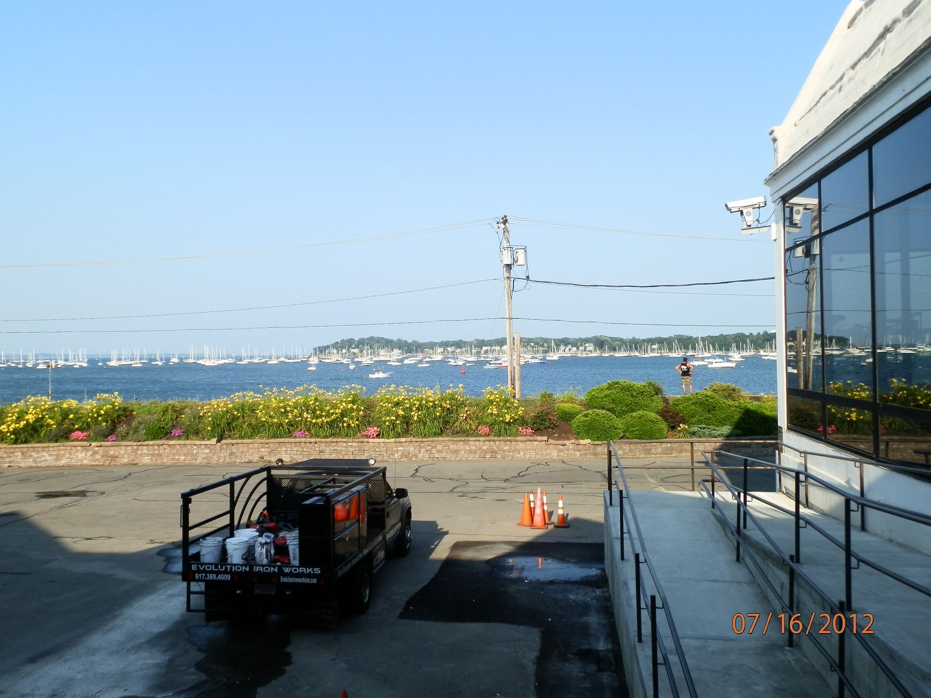 Riverside view from a parking lot with a truck, flowers, and a glass building under a clear blue sky