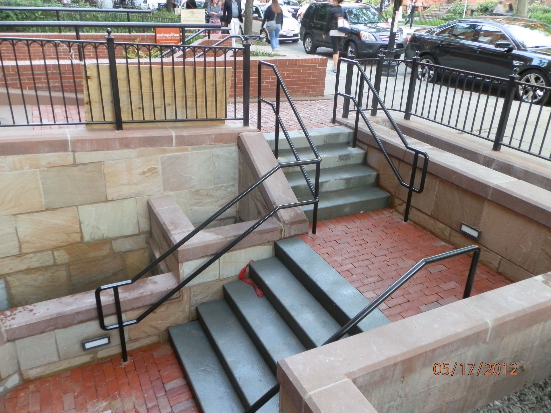 Steep outdoor stairs with black railings beside a stone retaining wall and sidewalk near parked cars.