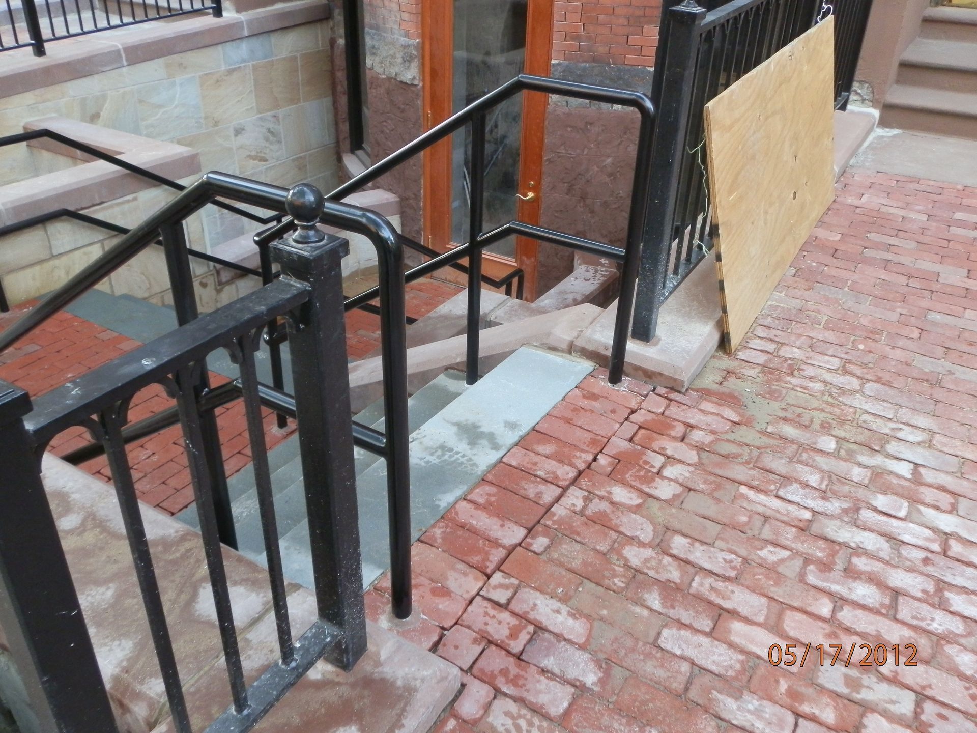 Ramps and black metal railings beside brick steps, with a plywood barrier on a narrow walkway.