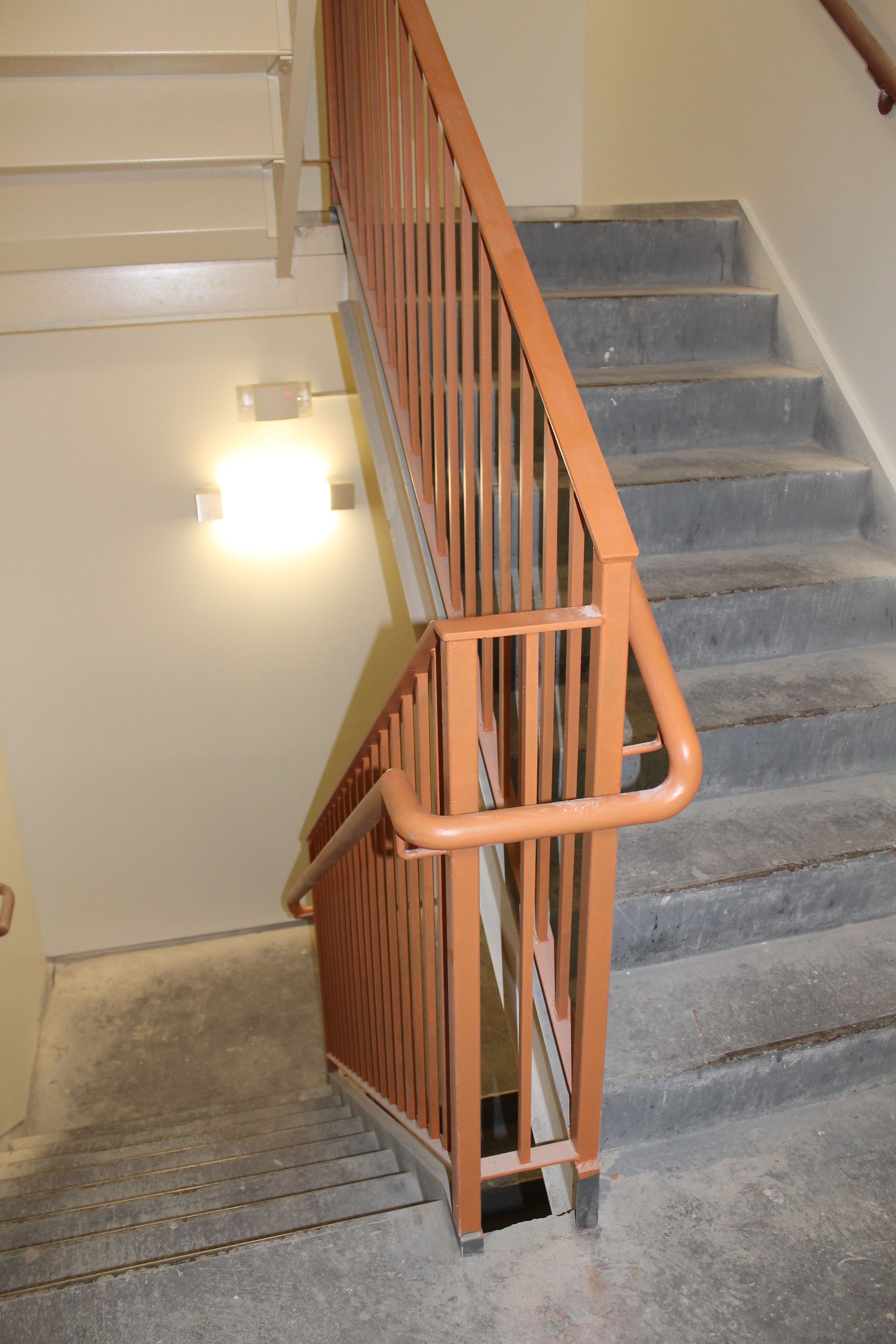 Stairwell with gray carpeted stairs, orange handrails, and a wall light.