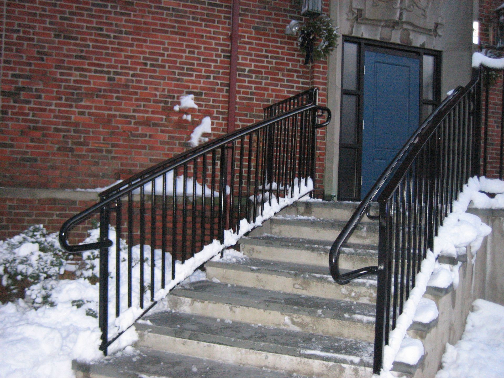 Snowy outdoor stairway with black railings leading to a blue door beside a brick wall