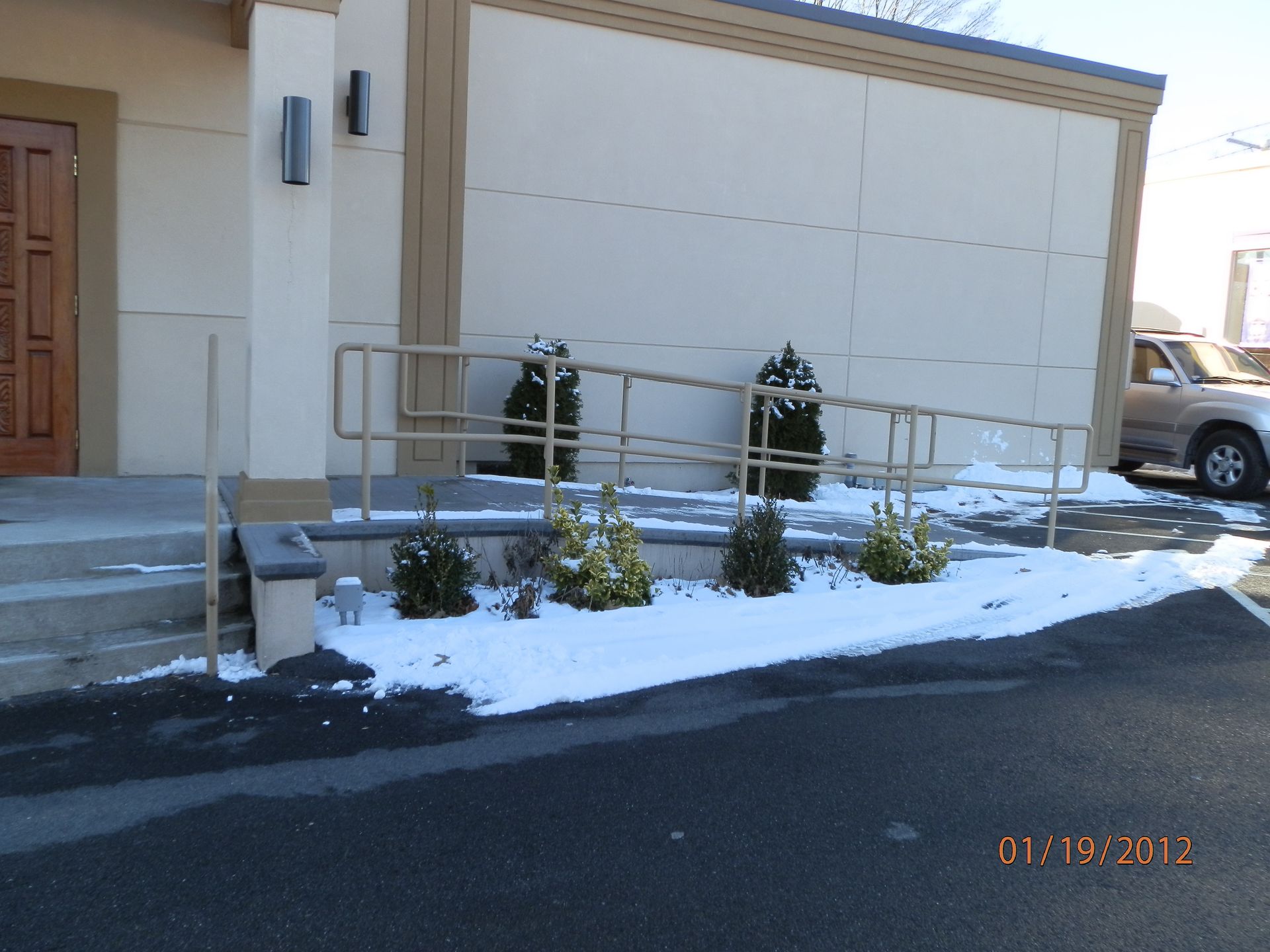 Snow-covered building entrance with steps, railing, shrubs, and a parked truck nearby