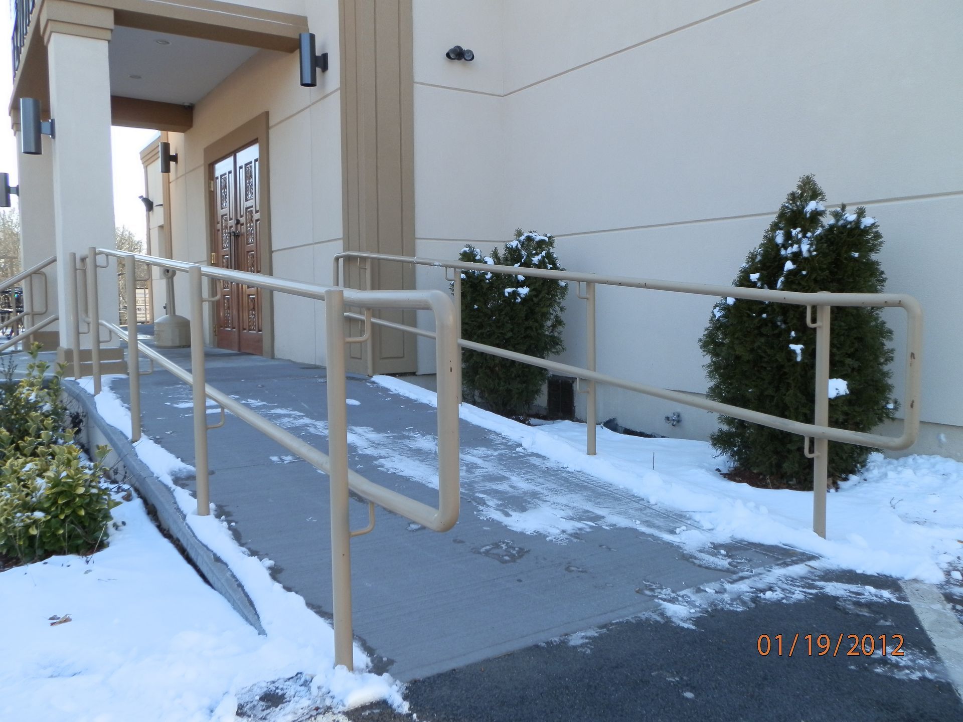 Snow-covered accessible ramp with beige handrails leading to a building entrance