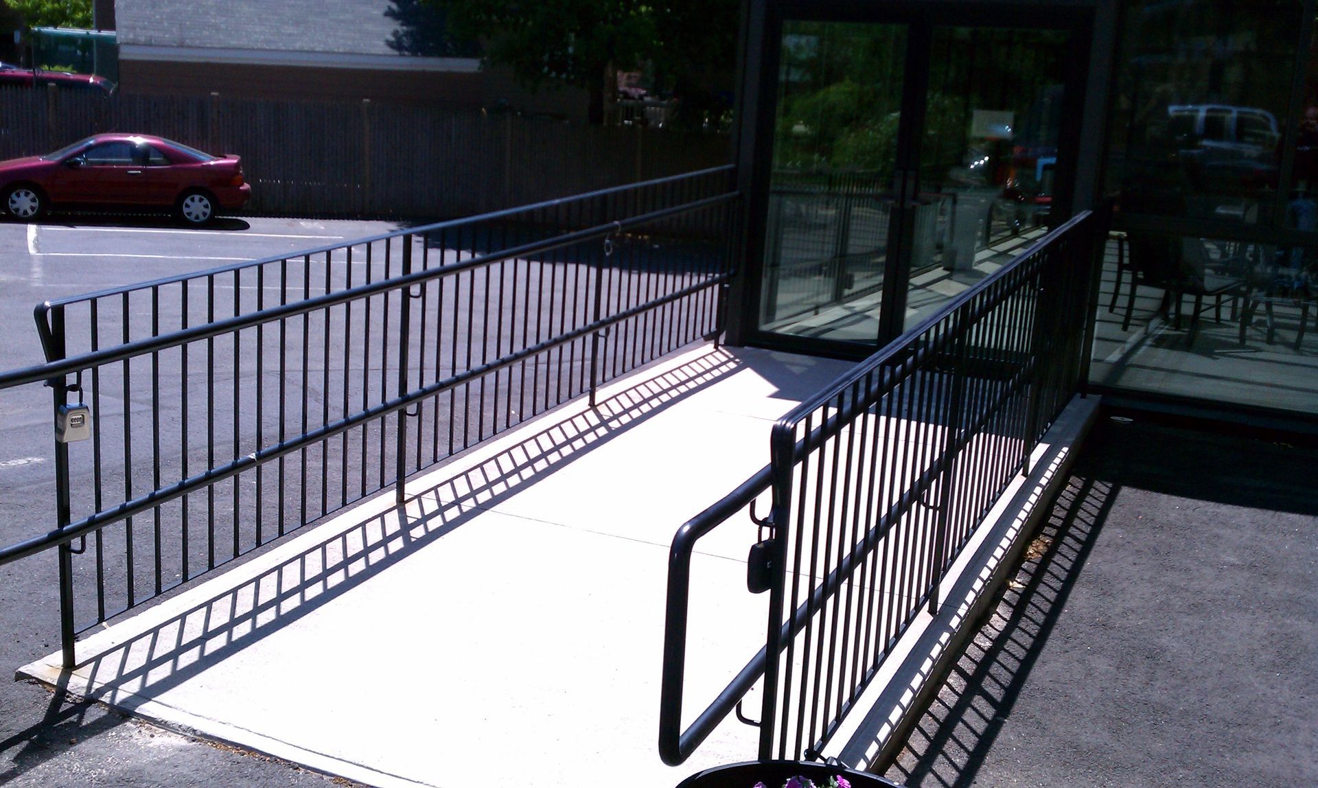 Ramped walkway with black railings leading to a glass entrance beside a street and parked car.