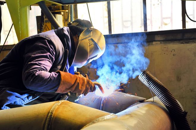 A person in a welding helmet and protective gloves works on a metal pipe, creating bright blue sparks and fumes.