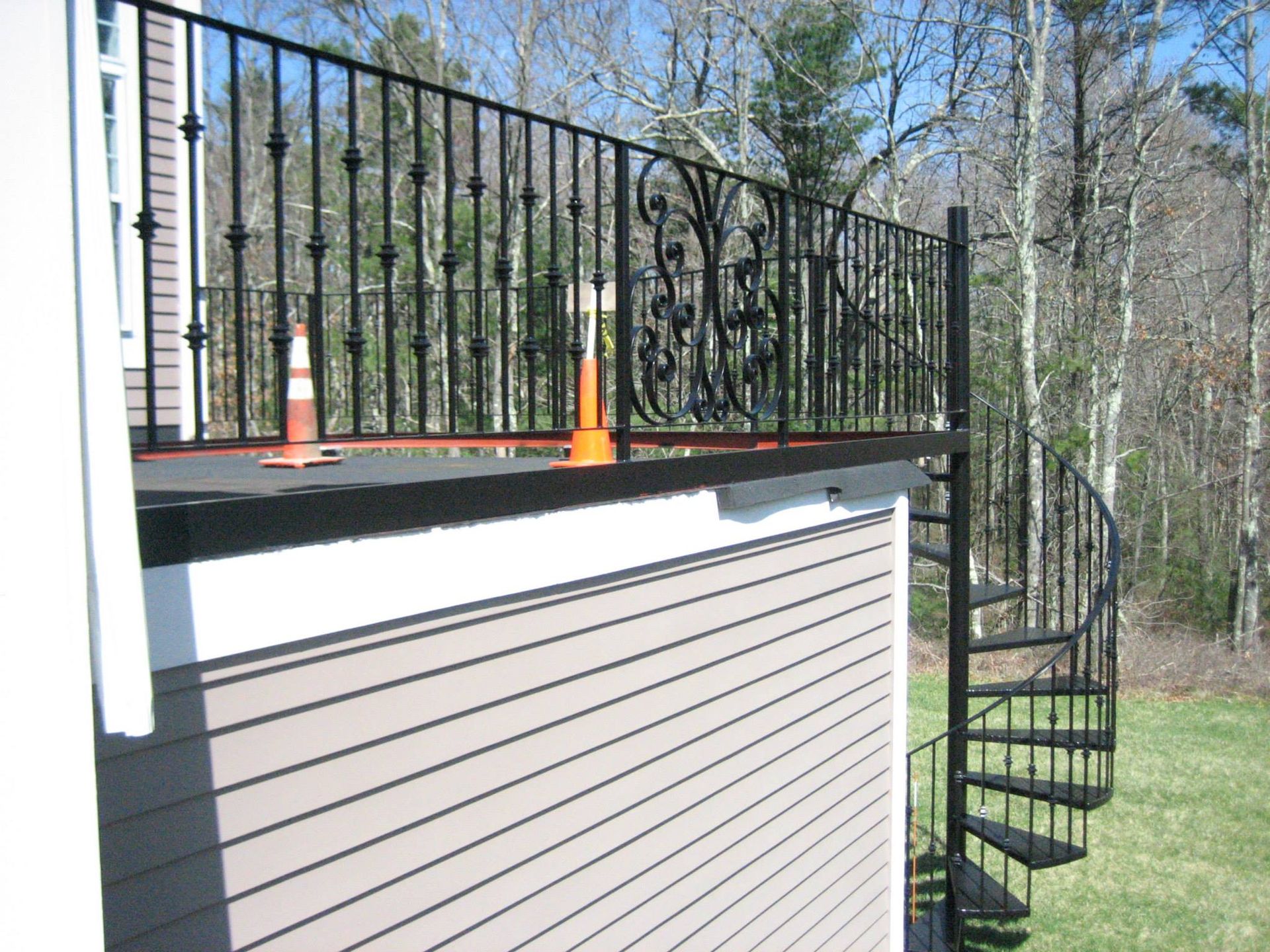 House exterior with deck railing, orange cones, and black spiral staircase beside a wooded yard