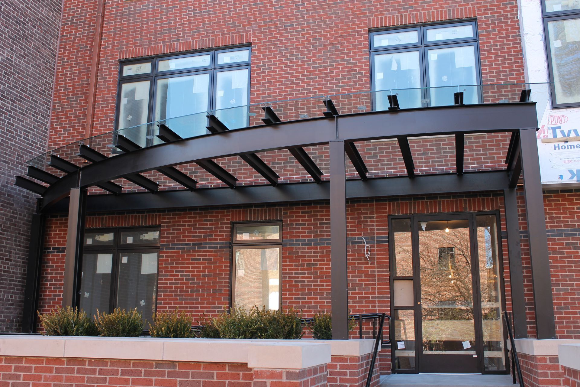 Brick building entrance with black metal awning, glass doors, and windows above a low brick wall.