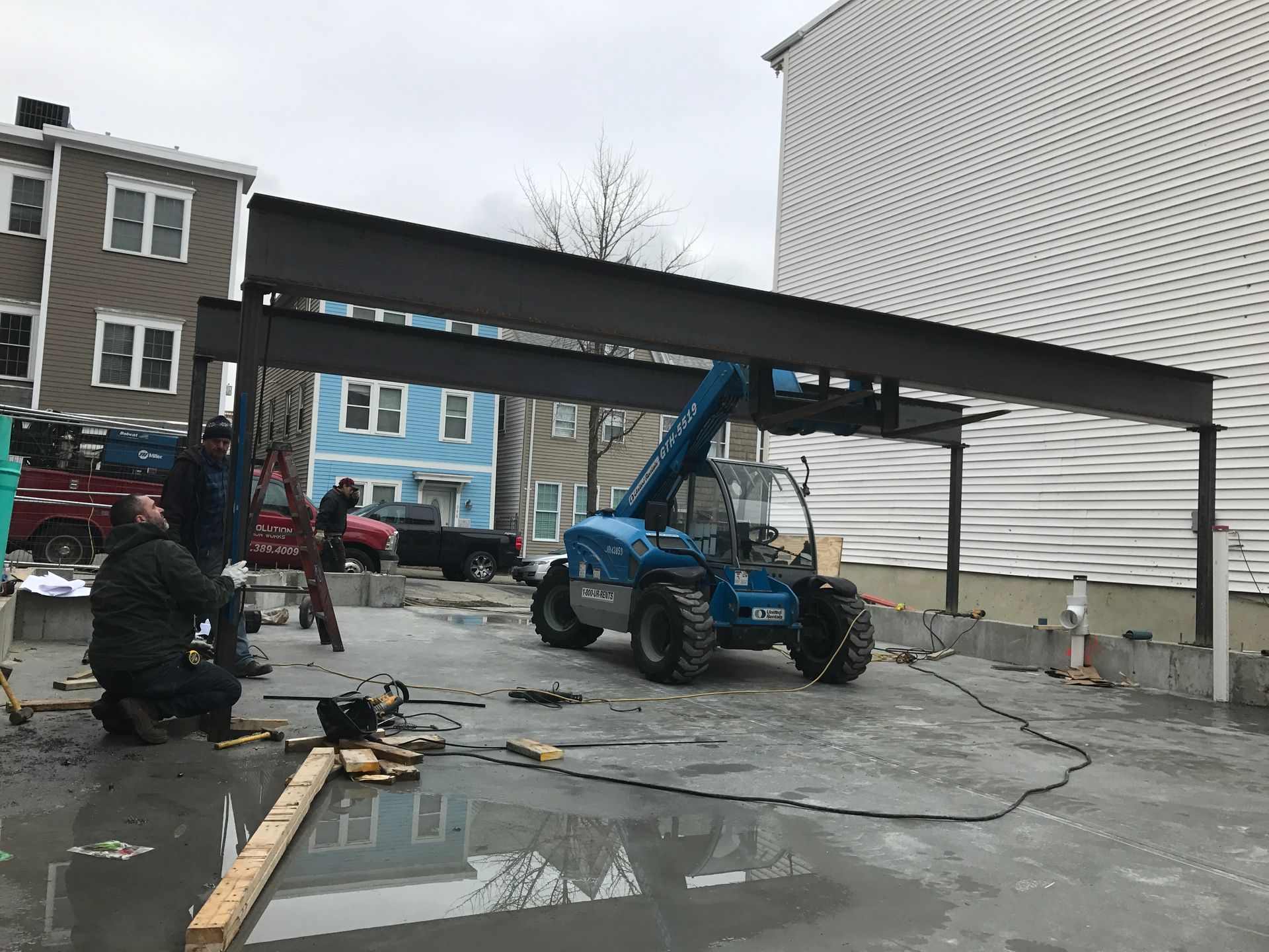 Blue forklift lifts debris in a wet alley beside damaged buildings after a storm.