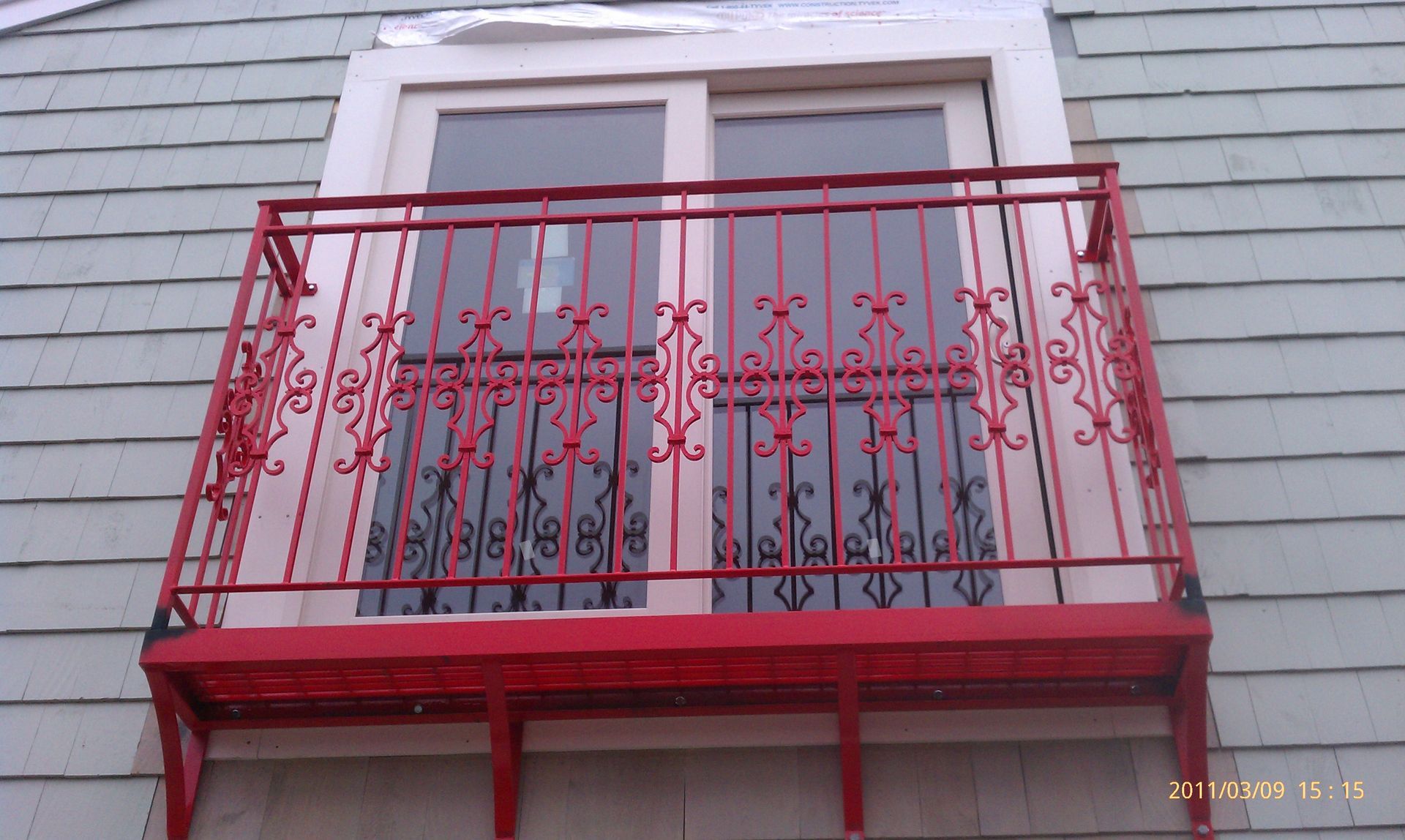 Red ornate balcony railing in front of a window on a gray house exterior