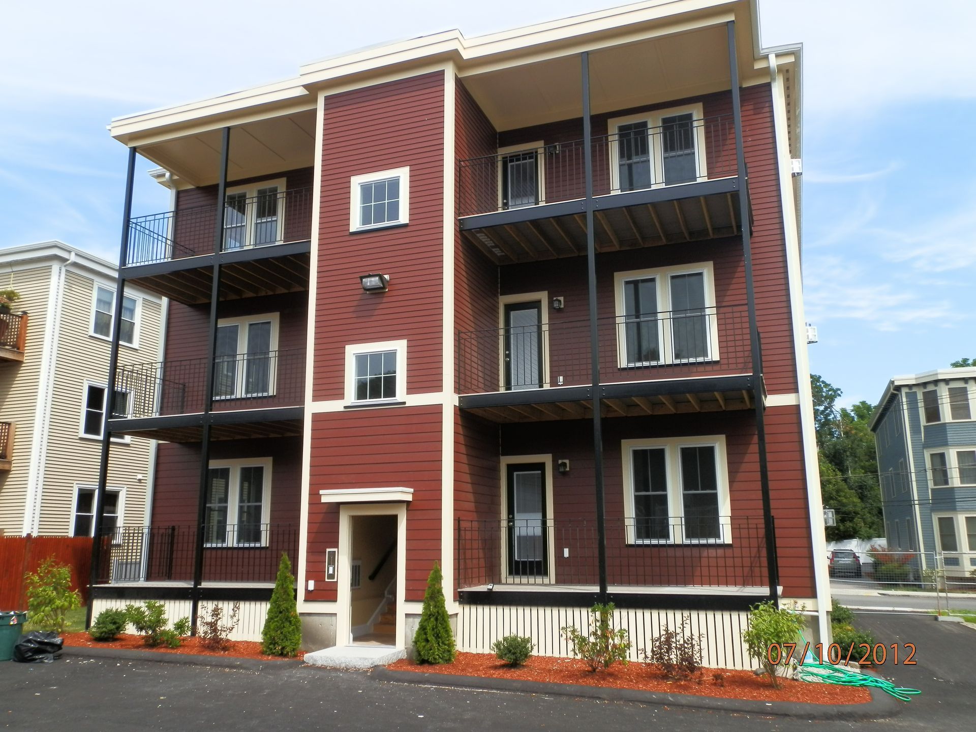 Three-story red apartment building with balconies and white trim under a blue sky