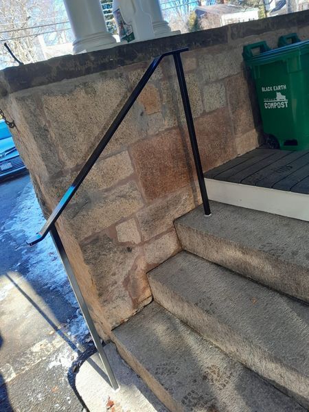 A black metal handrail installed on outdoor concrete steps next to a stone wall and a green trash bin.