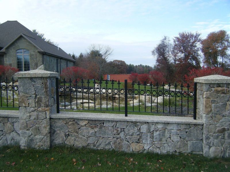 A stone wall with wrought iron fencing in front of a house, set against a backdrop of trees with autumn foliage.