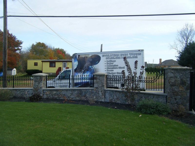 A utility truck parked behind a stone wall and metal fence on a grassy lawn with a yellow mobile home in the background.