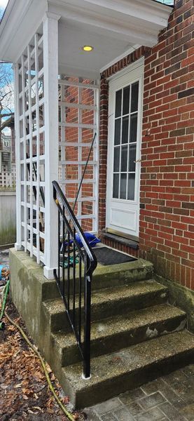 Concrete steps with a black metal railing leading to a white door under a white trellis-style porch cover.