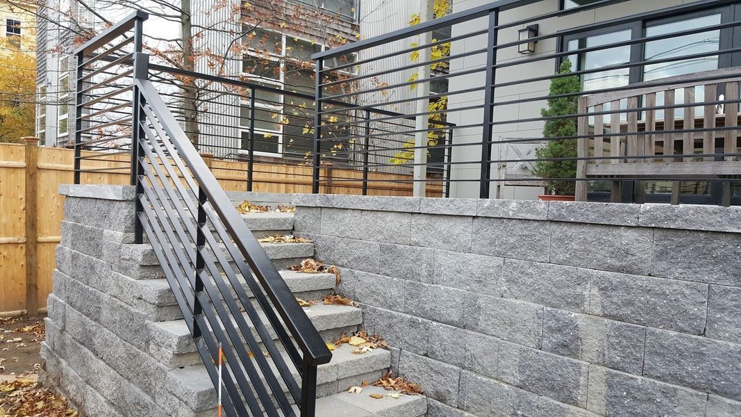 Stone stairs leading to a patio with a metal railing, framed by a stone retaining wall and wooden fence.