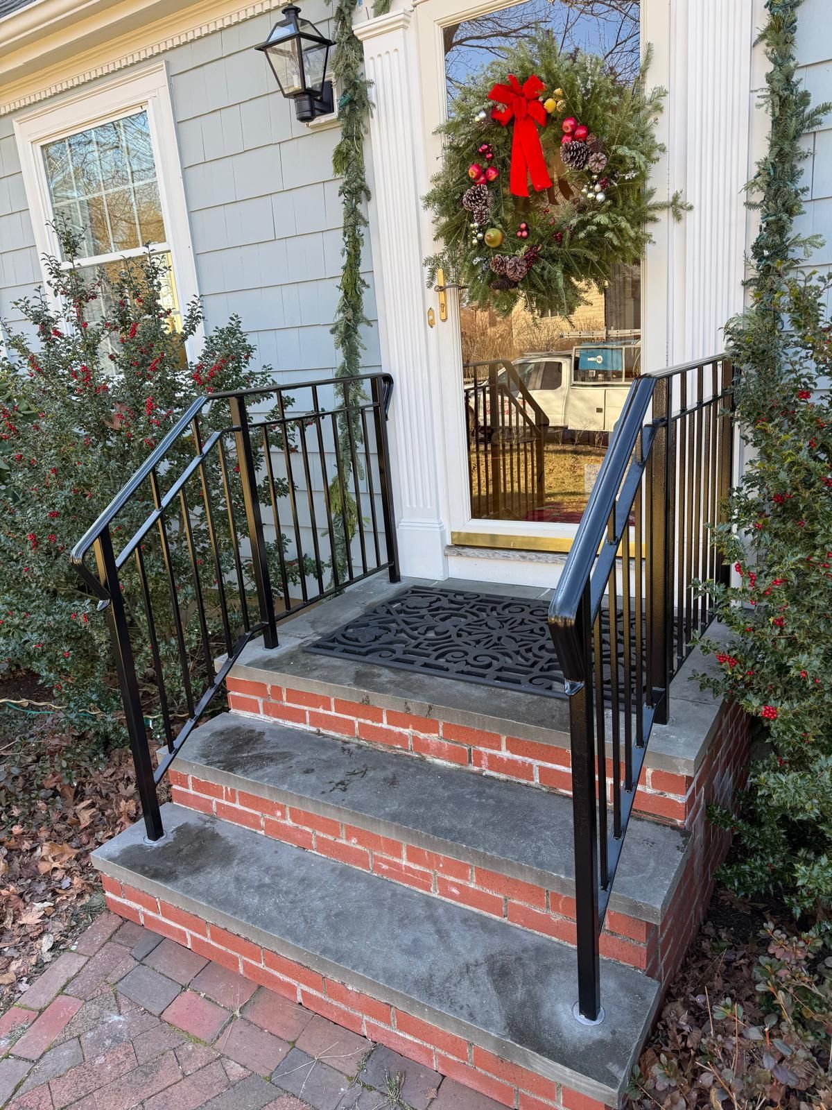 Front porch steps with black railings, a brick walkway, and a front door decorated with a holiday wreath
