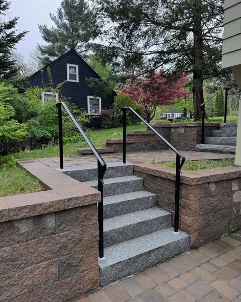 Outdoor concrete steps with black metal handrails set between retaining walls leading to a path and a black house.