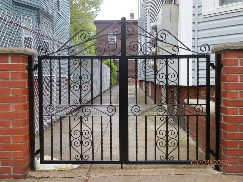 A black wrought iron double gate set between two red brick pillars at the entrance to a paved alleyway.