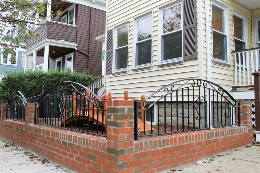 A decorative iron fence with arched panels atop a red brick wall, situated in front of a house.