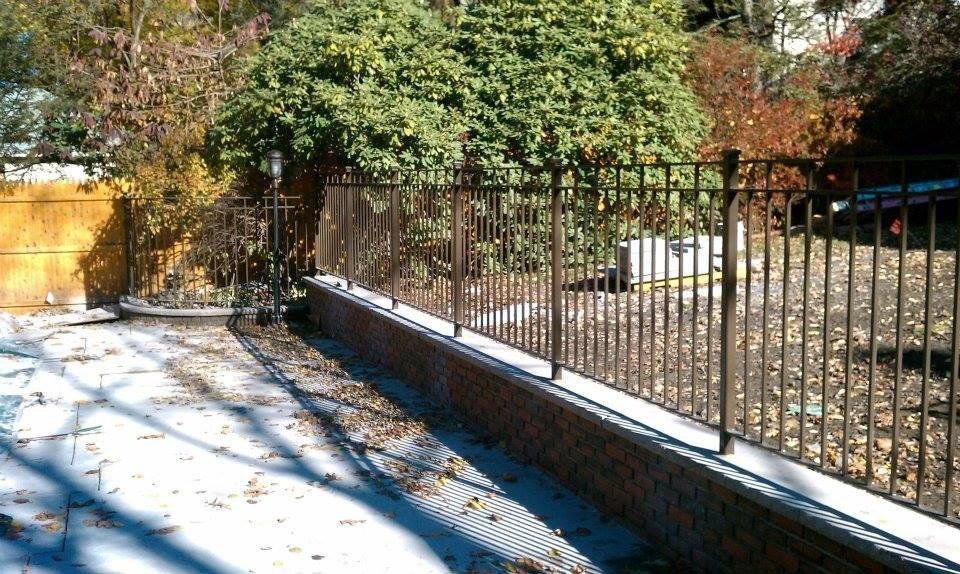 A metal fence sits atop a brick retaining wall next to a concrete pool deck covered in fallen leaves.