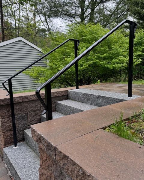 Black metal handrails installed on grey stone outdoor steps next to a red brick retaining wall.
