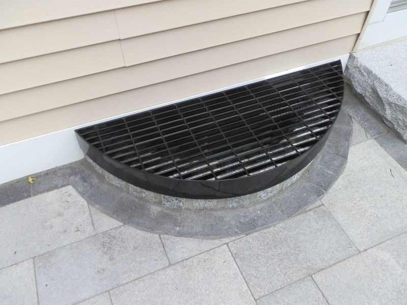 A black metal grate covering a semicircular window well set into the foundation of a house with beige siding.