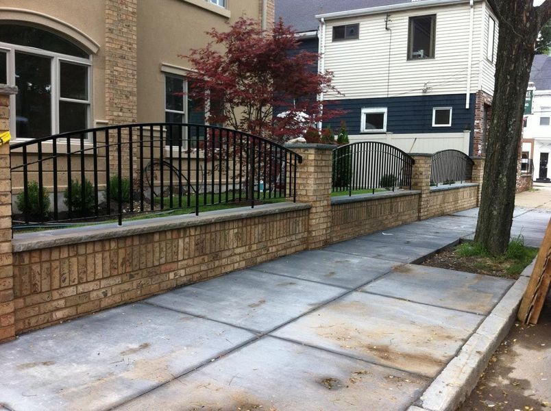 A tan brick wall topped with curved black metal fencing runs alongside a concrete sidewalk in front of a house.