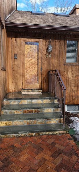 A wooden-sided house entrance with stone stairs, a wrought-iron railing, and a red brick walkway under a blue sky.
