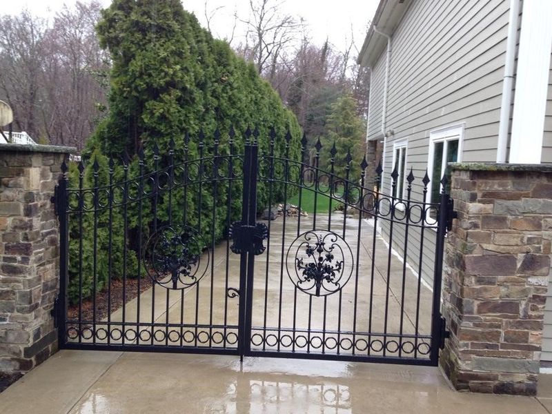 A double black iron gate with circular ornamental detail, installed between two stone pillars next to a house and hedges.